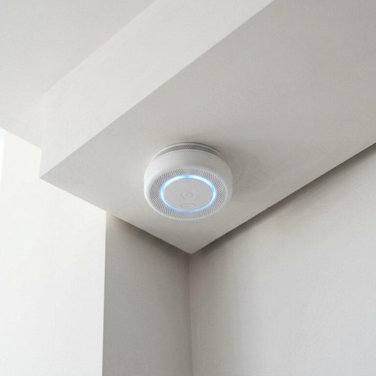A close-up of a modern smart smoke detector mounted on a white ceiling with subtle blue status light glowing.